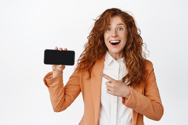 Gelukkig zakenvrouw demonstarter app wijzende vinger naar mobiele telefoon met opgewonden glimlach dragen pak staande over witte achtergrond