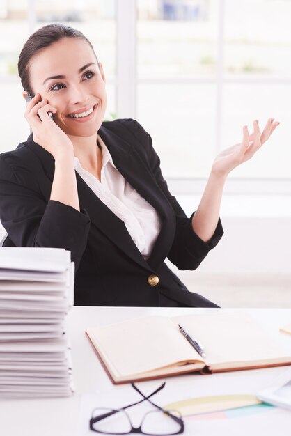 Gelukkig zakenvrouw. Gelukkige jonge vrouw in formalwear die op de mobiele telefoon praat terwijl ze op haar werkplek zit