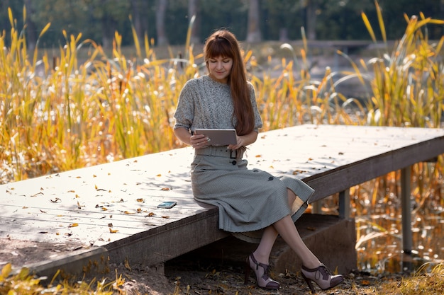 Gelukkig zittende vrouw in het park bij de vijver met een digitale tablet tijdens herfstwandelingen