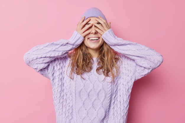 Gelukkig zorgeloze jonge vrouw bedekt ogen met handen probeert te verbergen voor iemand glimlacht breed draagt hoed en casual trui geïsoleerd over roze hebben. Positieve menselijke emoties en gevoelens concept