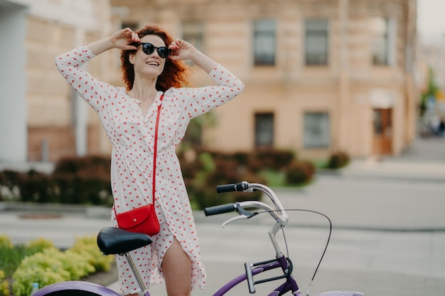 Gelukkig zorgeloze vrouw fietst in stad poses in de buurt van fiets houdt handen op tinten gekleed in witte jurk geniet van zomertijd en vakantie gericht opzij lege kopie ruimte over onscherpe stedelijke achtergrond