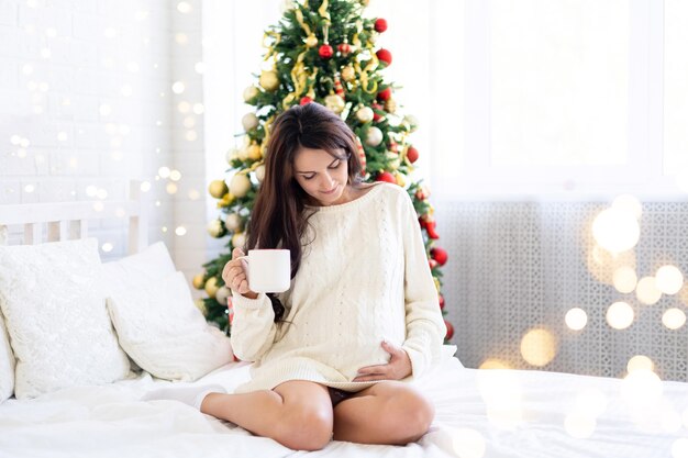 Gelukkig zwangere vrouw met kopje koffie of thee in bed thuis slaapkamer over kerstboom achtergrond