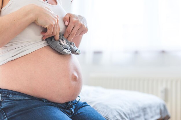 Gelukkig zwangerschapsconcept Moeder in spijkerbroek houdt de buik van babyschoenen vast op de onscherpe achtergrond