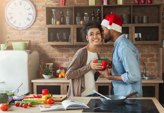 Gelukkig zwarte man met kerstmuts die zijn vrouw verrast met een cadeau in de keuken terwijl ze het avondeten kookt