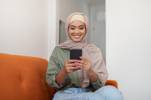 Gelukkig zwarte vrouw in hijab met behulp van telefoon om thuis te zitten