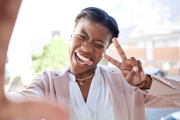 Gelukkig zwarte vrouw vredesteken en selfie in de stad met tong uit voor fotogeheugen of vlog buitenshuis Gezicht van vrouwelijke persoon of werknemer glimlach voor leuke foto humor of geluk in een stedelijke stad