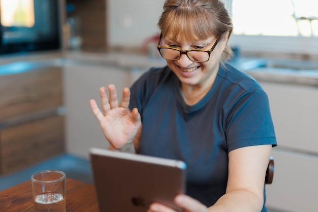 Gelukkige 50-jarige vrouw van middelbare leeftijd die thuis een digitale tablet gebruikt die in de keuken zit