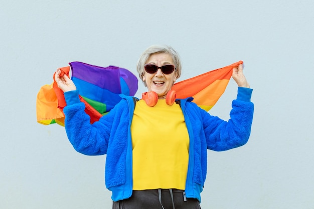 Gelukkige 80-jarige vrouw, kleurrijk gekleed in een zonnebril en trots zwaaiend met de regenboogvlag van de homogemeenschap