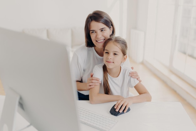 Gelukkige aanhankelijke moeder omarmt haar kleine dochter, werk samen aan moderne computer, preapare presentatie voor les kijk graag naar monitor breng vrije tijd thuis door surf internetpagina