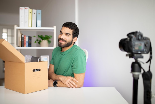 Gelukkige aantrekkelijke duizendjarige Arabische man met baard op de werkplek met camera en kartonnen doos