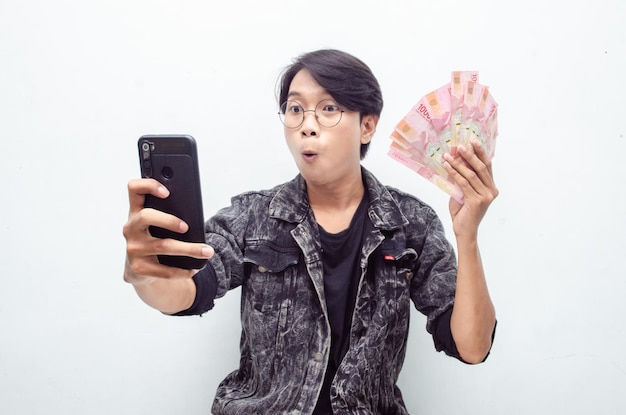 Gelukkige aantrekkelijke indonesische jongeman schokte gelukkig terwijl hij rupiah-papiergeld en telefoon vasthield.