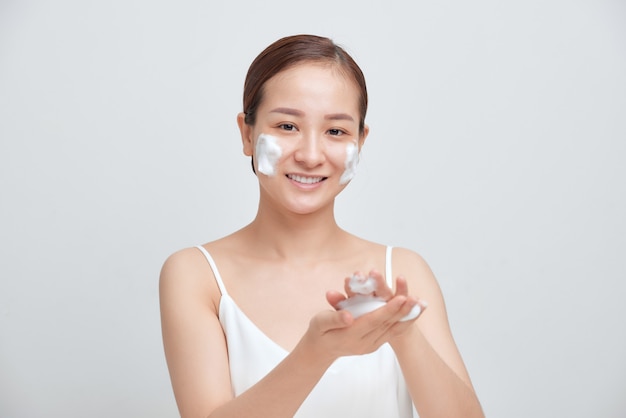 Gelukkige aantrekkelijke jonge Aziatische vrouw die schuimend reinigingsmiddel op haar gezicht op witte achtergrond toepast.