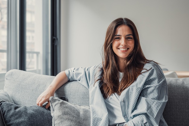 Gelukkige aantrekkelijke jonge volwassen vrouw meisje zit op de bank en kijkt naar de camera in een modern gezellig huis glimlachende alleenstaande vrouw van twintig jaar mooi gezicht ontspannen op de bank alleen genieten van welzijn poseren voor portretxA