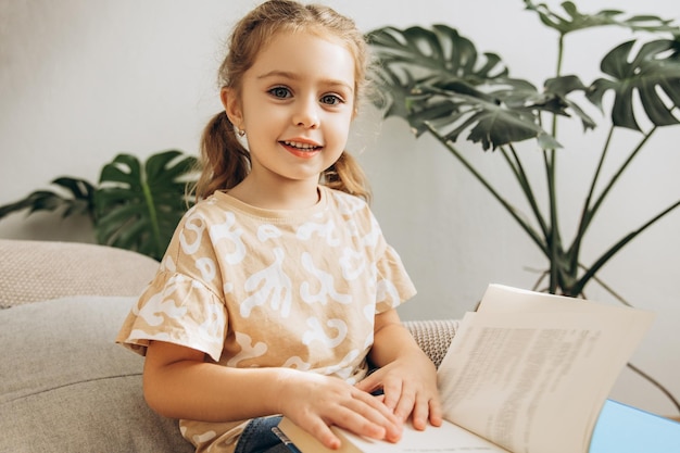 Gelukkige aantrekkelijke kleine meisje met een leesboek die naar de camera kijkt en op een comfortabele bank zit