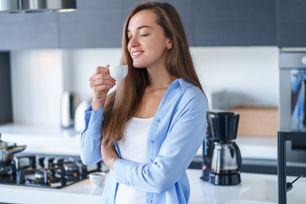 Gelukkige aantrekkelijke vrouw die hete verse aromatische koffie drinken en thuis van koffiearoma in de keuken genieten