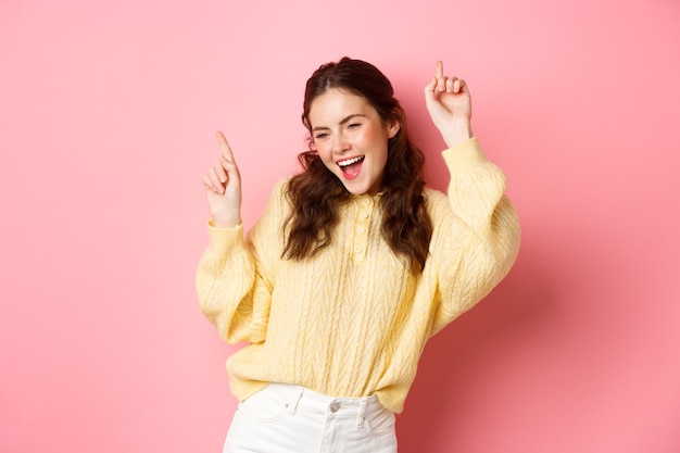 Gelukkige aantrekkelijke vrouw die pret heeft, vingers omhoog wijst en zorgeloos danst, vrolijk glimlachend, staand tegen roze muur.