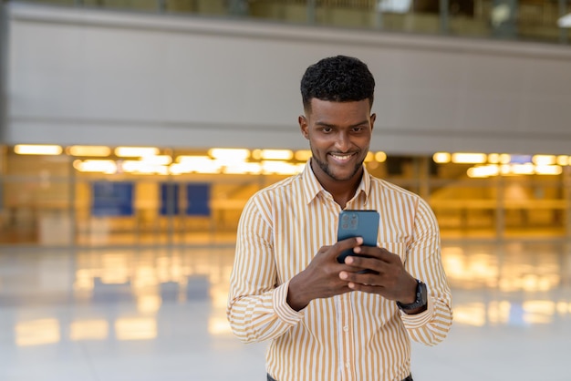 Gelukkige Afrikaan op luchthaventerminal die tijdens covid reist terwijl hij lacht en mobiele telefoon gebruikt