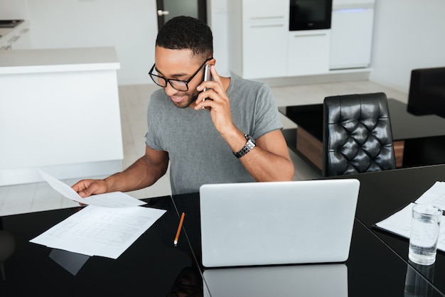 Gelukkige afrikaanse man gekleed in grijs t-shirt en bril die op mobiele telefoon praat terwijl hij laptop gebruikt en op papieren kijkt.