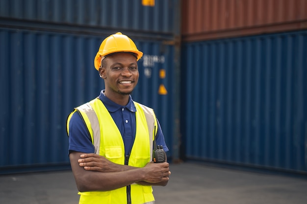 Gelukkige Afrikaanse werknemer smailt, staat op de containerwerkplek en gekruiste armen met een gevoel van geluk