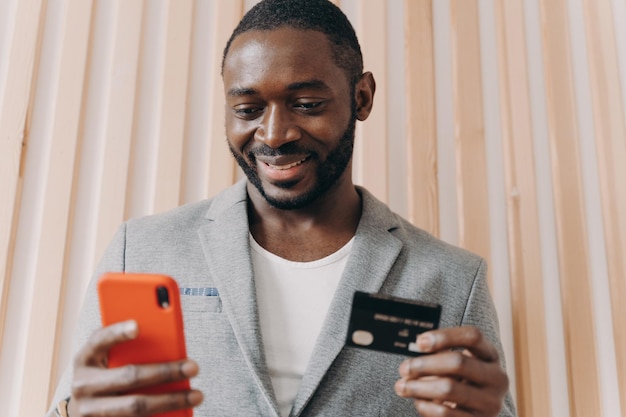 Gelukkige Afrikaanse zakenman in pak met creditcard en mobiele telefoon terwijl hij online winkelt