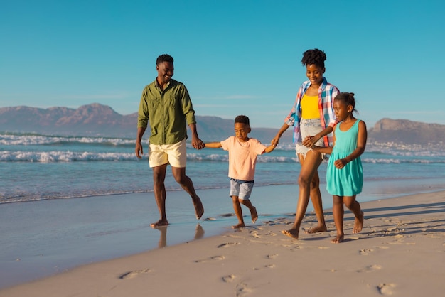 Gelukkige Afro-Amerikaanse jonge ouders houden de handen van kinderen vast en lopen op het strand tegen heldere hemel