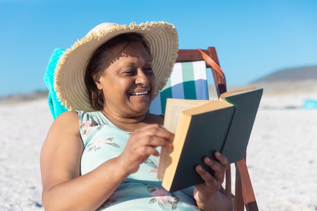 Gelukkige Afro-Amerikaanse senior vrouw die een boek leest terwijl ze op een klapstoel op het strand zit