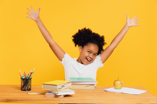 Gelukkige Afro-Amerikaanse vrouwelijke schoolmeisje zit aan het bureau en strekt haar arm omhoog tijdens de les