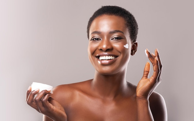 Gelukkige Afro-vrouw houdt en past vochtinbrengende crème op haar gezicht aan