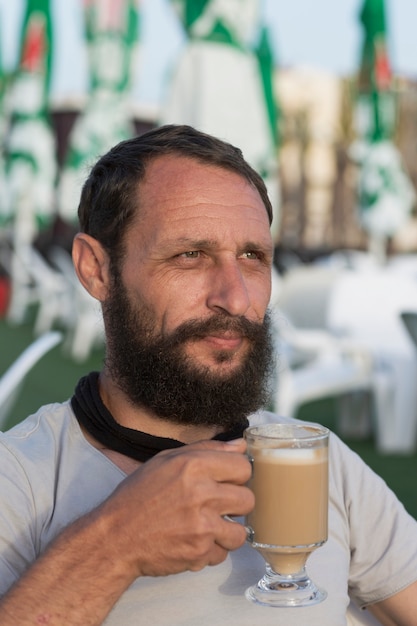 Gelukkige Amerikaanse kerel die van heerlijke kop koffie geniet. Portret van een knappe bebaarde man die hete espressokoffie drinkt met melk kijkt in de verte In zonsondergangverlichting. Man Met Een Koffiemok.