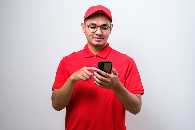 Gelukkige Aziatische bezorger die een bril draagt en op een mobiele telefoon wijst op een witte achtergrond