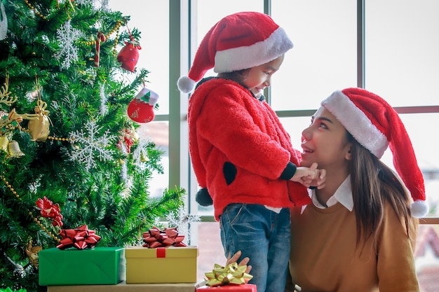 Gelukkige Aziatische familie moeder en dochter in rode en witte kerstman hoed en trui glimlachend samen spelen in de buurt van kerstboom met glitter decoratie artikelen en geschenkdozen in kerstavond.