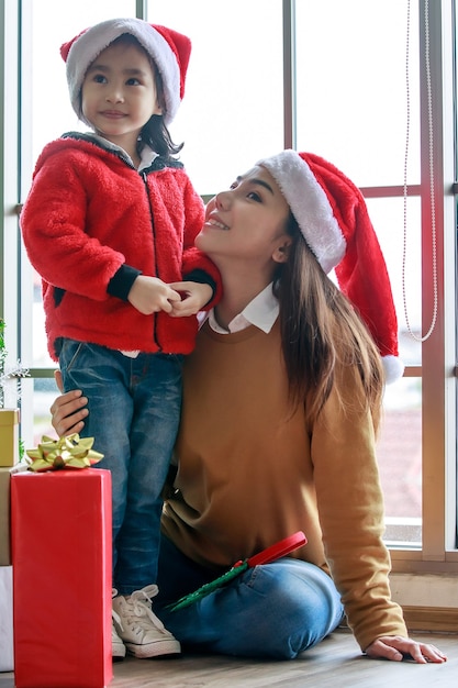 Gelukkige Aziatische familie moeder en dochter in rode en witte kerstman hoed en trui glimlachend samen spelen in de buurt van kerstboom met glitter decoratie artikelen en geschenkdozen in kerstavond.