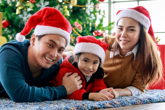 Gelukkige Aziatische familie vader moeder en dochter draagt een trui met rode en witte kerstman hoed op tapijtvloer vieren kerstavond samen voor decor kerstboom.
