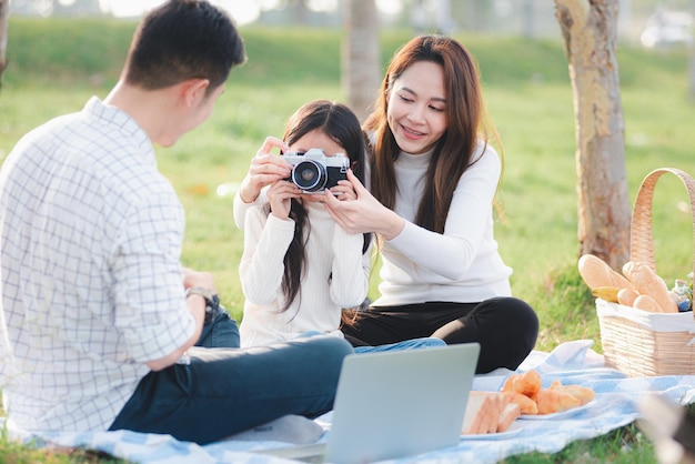 Gelukkige Aziatische jonge familie vader, moeder en kind hebben plezier en genieten van samen buiten zitten op het grasfeestje met foto's maken met een retro camera een picknick in het tuinpark op een zonnige dag