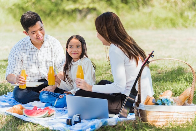 Gelukkige Aziatische jonge familie vader, moeder en kind klein meisje met plezier en genieten van buiten zitten op picknick deken drinken sinaasappelsap uit glazen fles, zomer rusten in een natuur tuin park