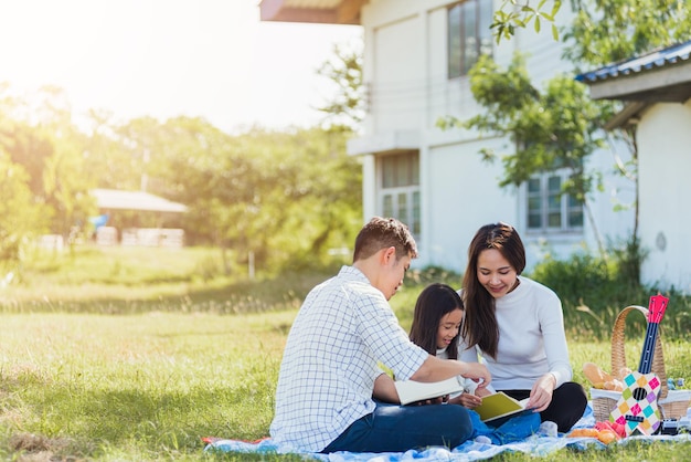 Gelukkige Aziatische jonge familie vader, moeder en kind meisje plezier hebben en genieten van buiten op picknick deken leesboek in park op zonnige tijd, zomer vrije tijd lente concept