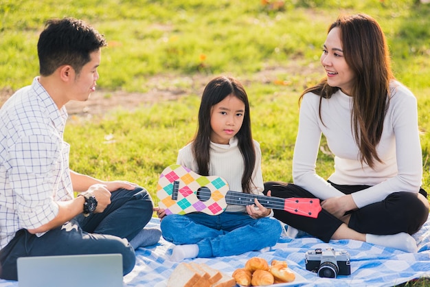 Gelukkige Aziatische jonge familie vader, moeder en kinderen die plezier hebben en samen buiten genieten zittend op het grasfeestje met Ukelele spelen tijdens een picknick in het tuinpark op een zonnige dag