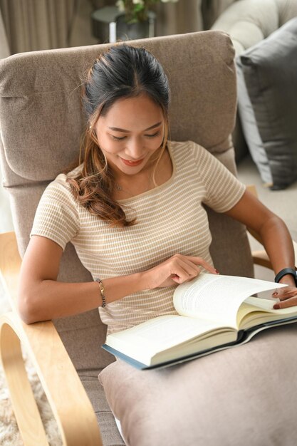 Gelukkige aziatische jonge vrouw ontspannen en chillen terwijl ze een boek of roman leest op haar comfortabele fauteuil thuis