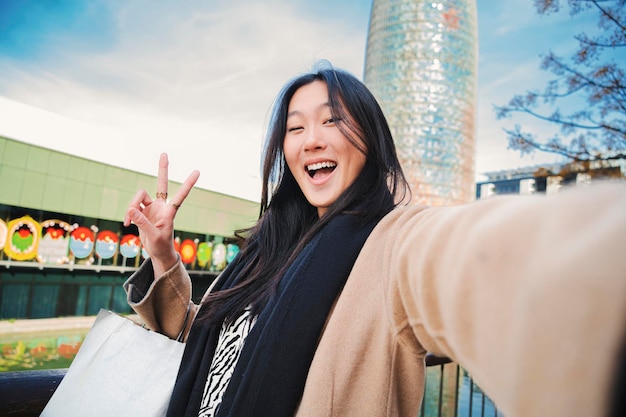 Gelukkige aziatische jongedame die een selfie-foto maakt met een smartphone Zelfportret van een Chinese lachende vrouw die het vredesteken doet na buitenshuis winkelen