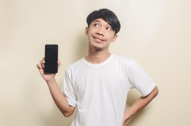 Gelukkige aziatische man die een helder wit t-shirt draagt met een gebaar dat het telefoonscherm op een geïsoleerde achtergrond toont