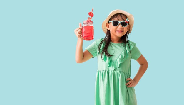 Gelukkige Aziatische meisje poseert met een hoed en zonnebril met een glas rood zoet water Vakantie zomer mode groene jurk geïsoleerd op pastelblauwe achtergrond