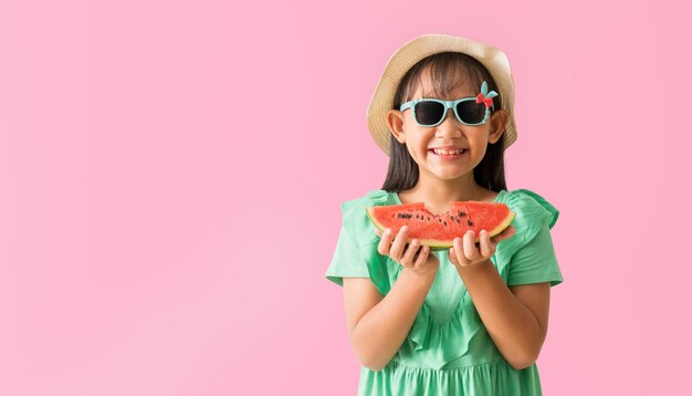 Gelukkige Aziatische meisje poseert met een hoed met een zonnebril die watermeloensnijden vasthoudt Vakantie zomer mode groene jurk geïsoleerd op pastel roze achtergrond