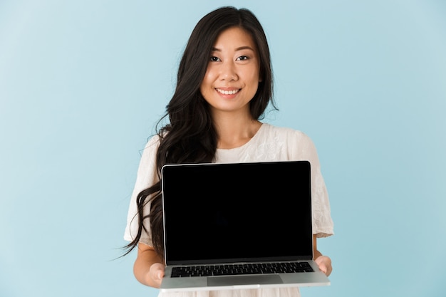 Gelukkige Aziatische mooie vrouw die over blauwe muur wordt geïsoleerd die vertoning van laptop toont