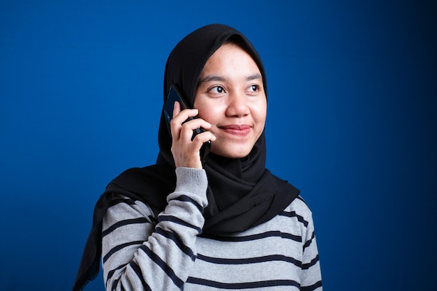 Gelukkige Aziatische moslimvrouw die hijabgesprekken op telefoon draagt, wijfje dat slimme telefoon gebruikt, communicatieconcept, tegen blauwe achtergrond