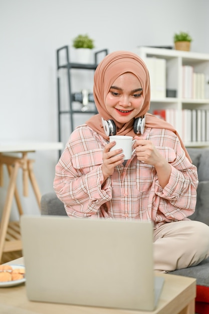 Gelukkige Aziatische moslimvrouw die koffie drinkt terwijl ze naar een film kijkt op haar laptop in de woonkamer