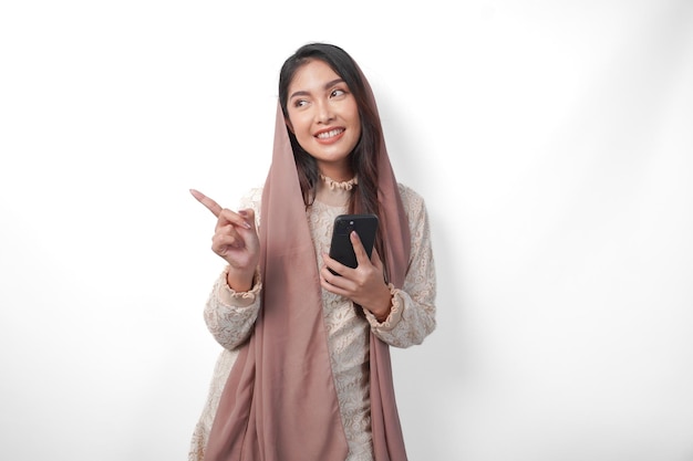 Gelukkige Aziatische moslimvrouw in sluier hijab die een smartphone vasthoudt terwijl ze wijst naar de kopieerruimte aan de zijkant over een geïsoleerde witte achtergrond Ramadan concept