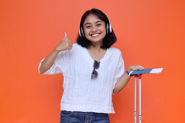 Gelukkige Aziatische reizigersvrouw luistert naar muziek terwijl ze duim opgeeft over oranje achtergrond