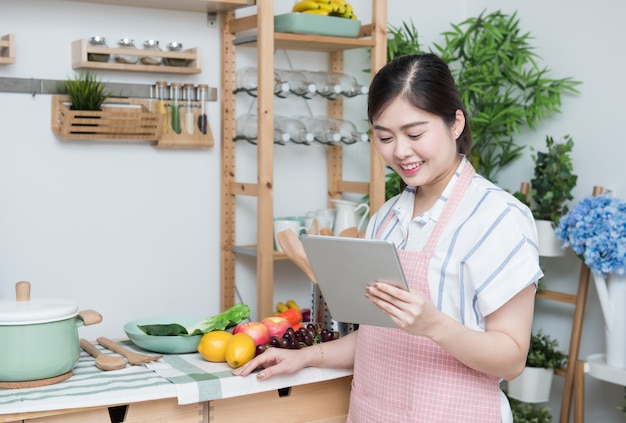 Gelukkige Aziatische vrouw die de keukenlezing van de receptentablet thuis het koken kijken