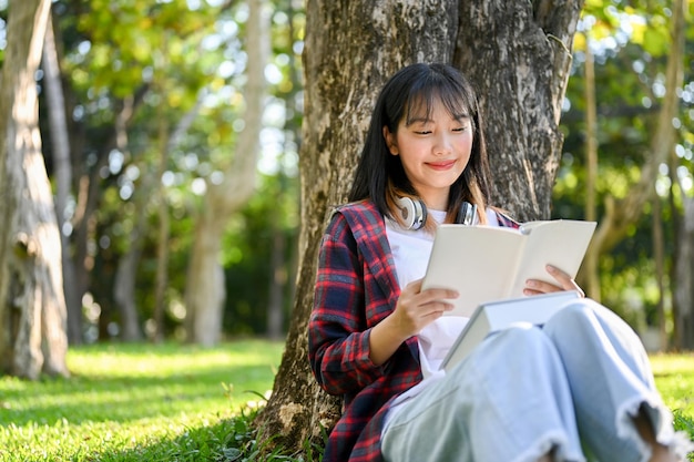 Gelukkige Aziatische vrouw die een boek of een roman leest onder de boom in het prachtige groene park