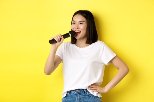 Gelukkige aziatische vrouw die lied zingt in karaoke, microfoon vasthoudt en opzij kijkt met een vrolijke glimlach, staande over gele achtergrond.